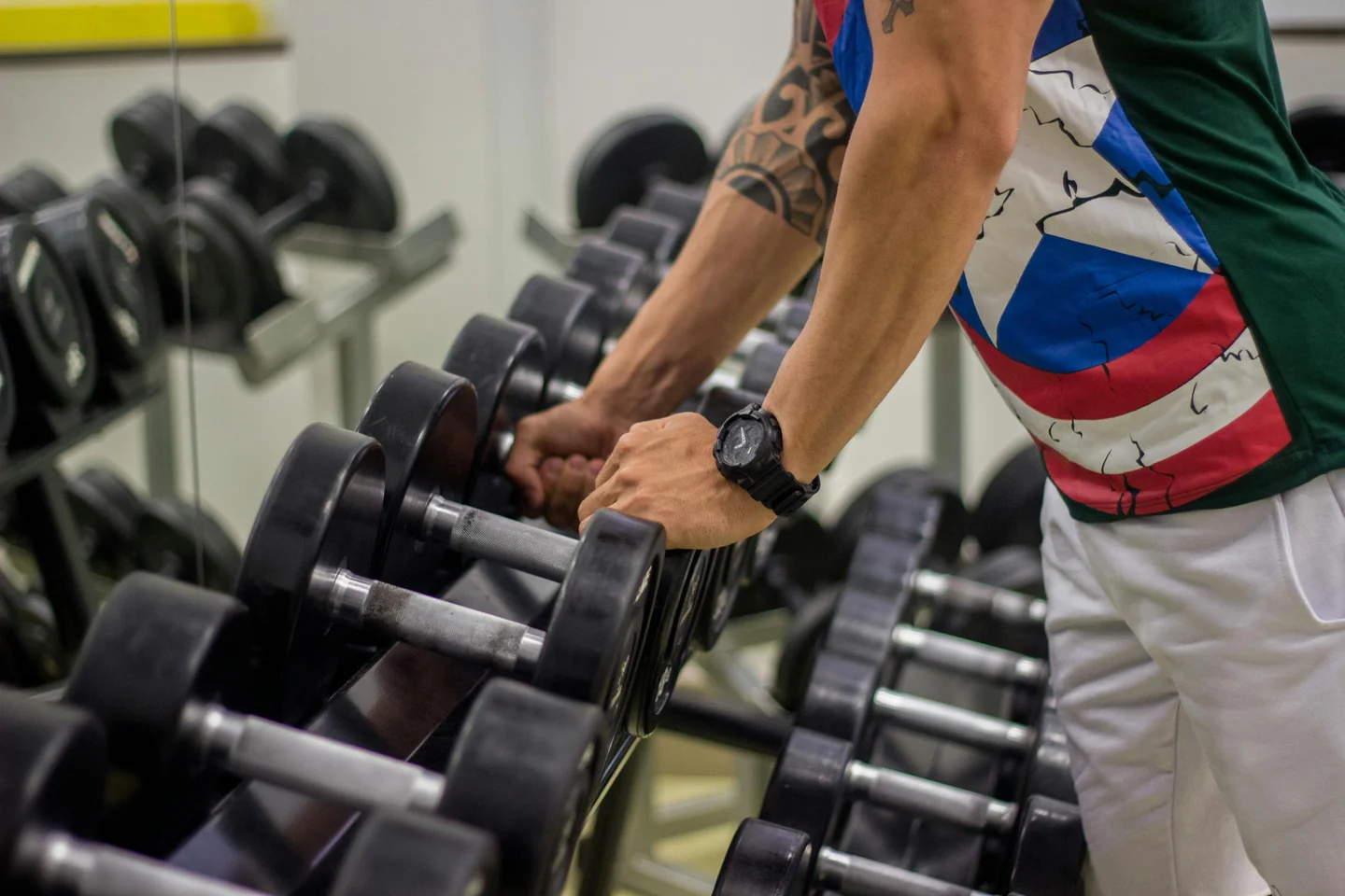 A lifter's tattooed hand picking up a dumbbell from a rack