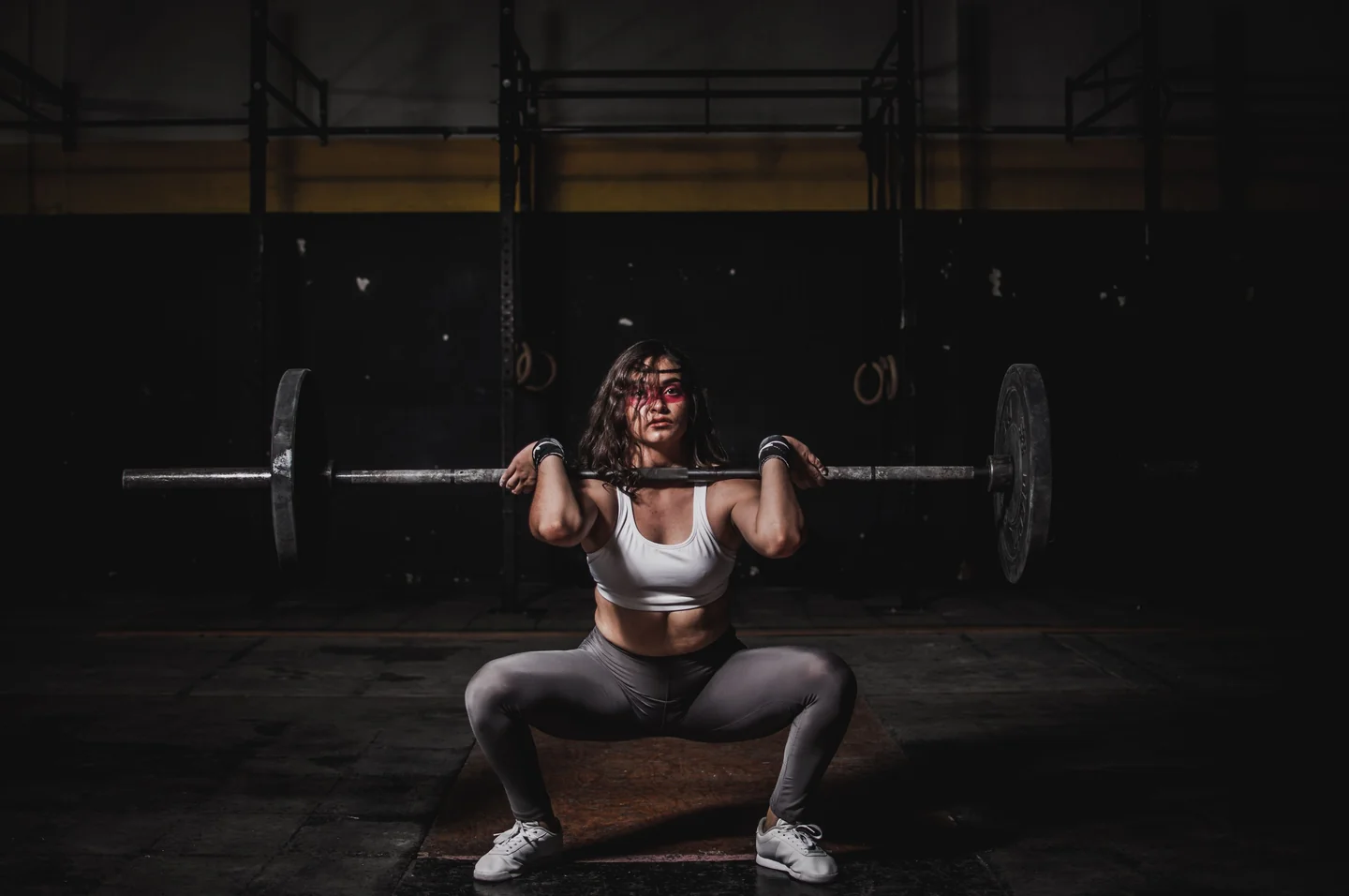 Una mujer en una posición de sentadilla frontal profunda con una barra en un gimnasio oscuro.