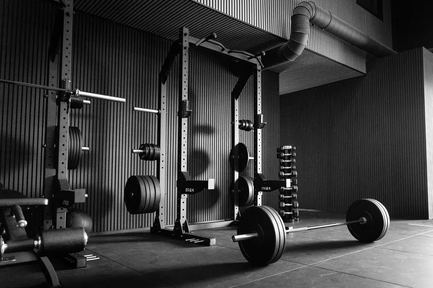 Una barra cargada apoyada en el suelo frente a jaulas de potencia vacías en un gimnasio tranquilo